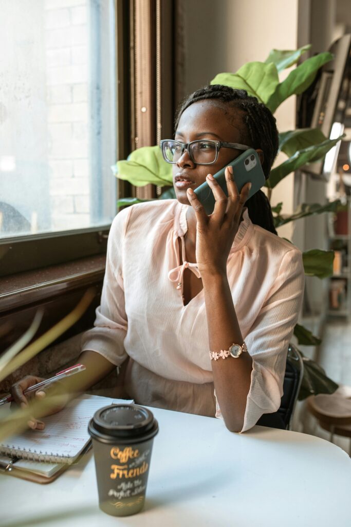 pexels-photo-5915256-5915256 Young woman with eyeglasses talking on smartphone at cafe table with coffee, notebook, and plants.